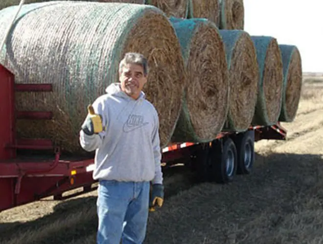A man standing next to a trailer with bales of hay.